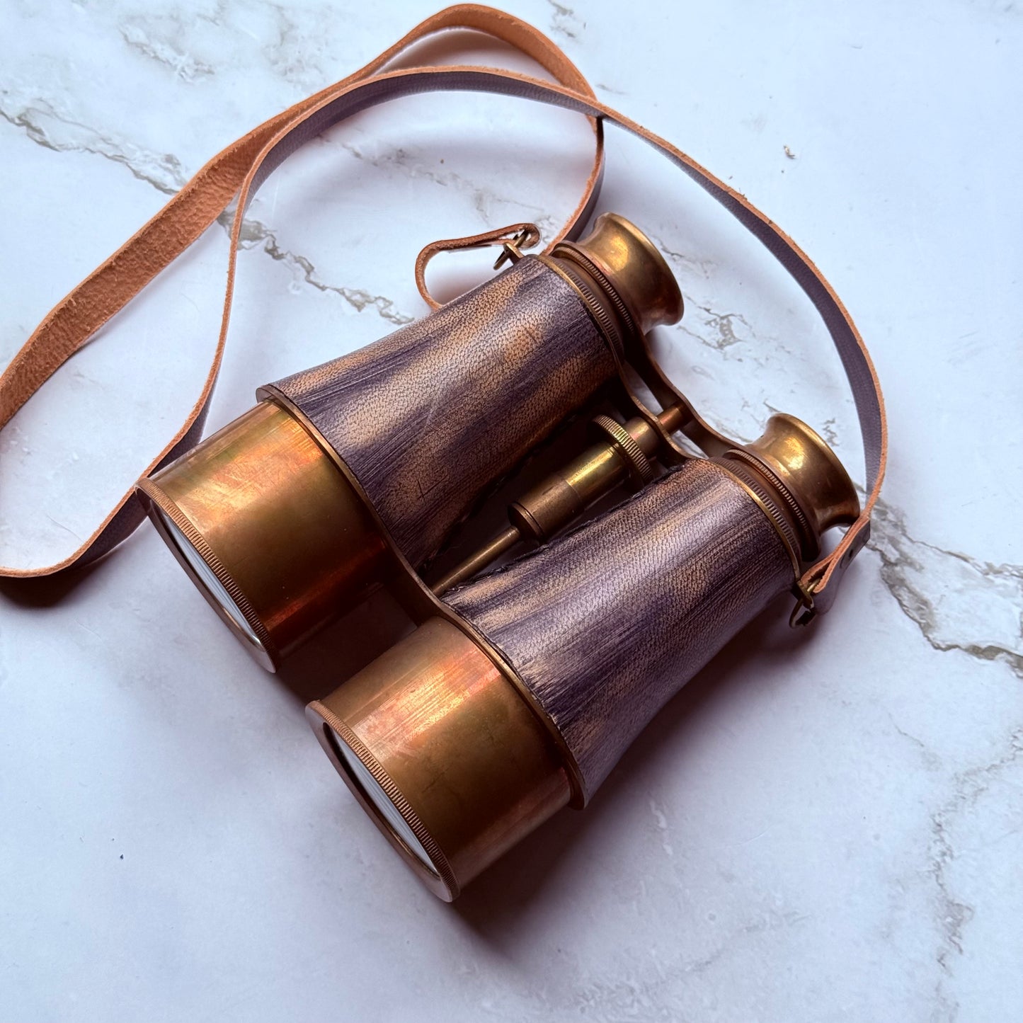 Vintage-style brass binoculars with leather strap on a marble surface