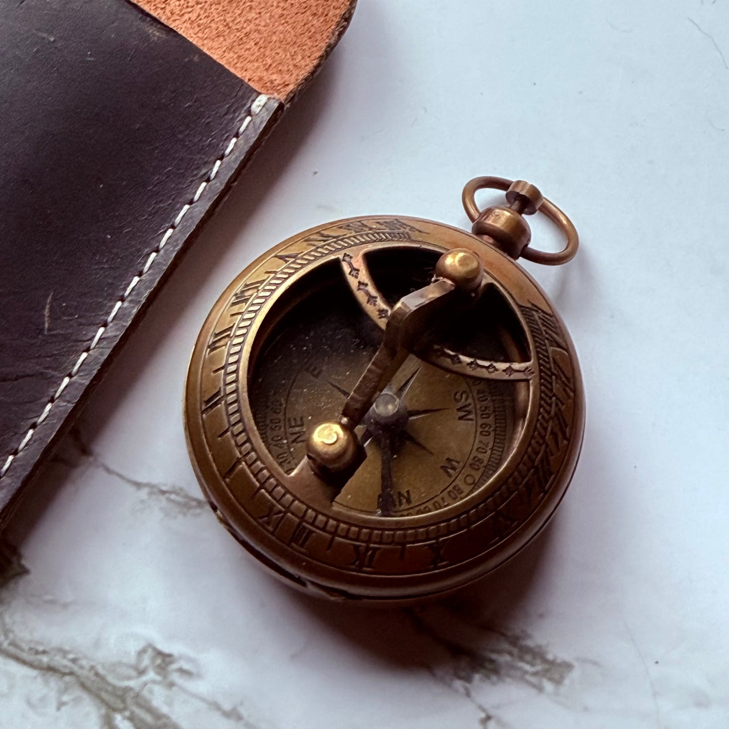 Brown leather case with a bronze pocket sundial compass on a white marble surface
