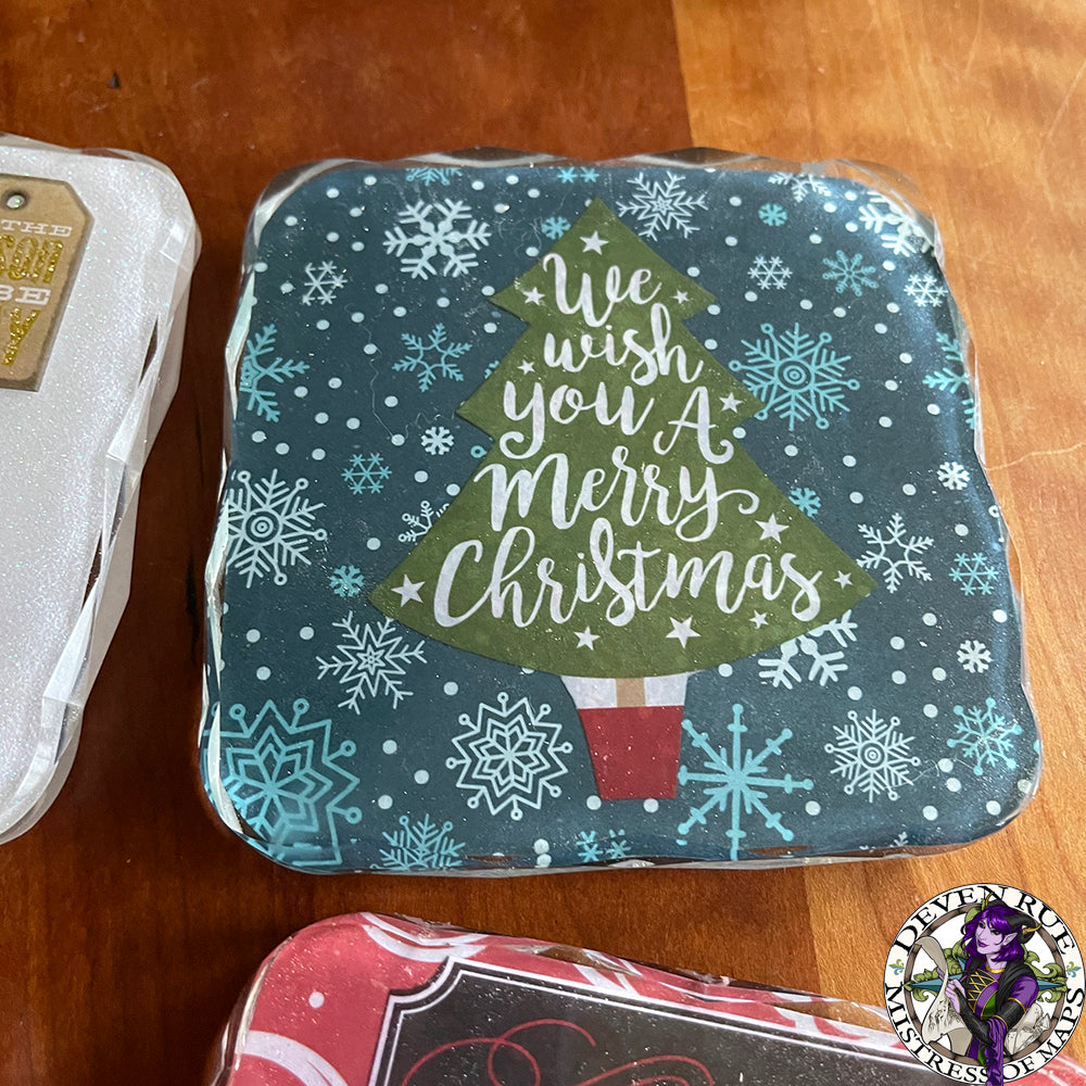 A close up of a resin coaster with blue background, snowflakes, and a Christmas tree with the words "We wish you a merry Christmas" written inside.