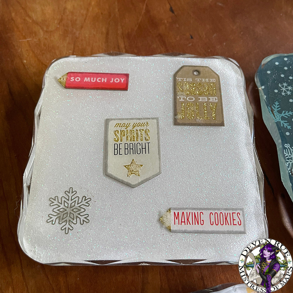 A close up of a coaster with a white sparkly background and tags saying "So much joy", "Tis the season to be jolly", "May your spirits be bright", and "Making cookies".