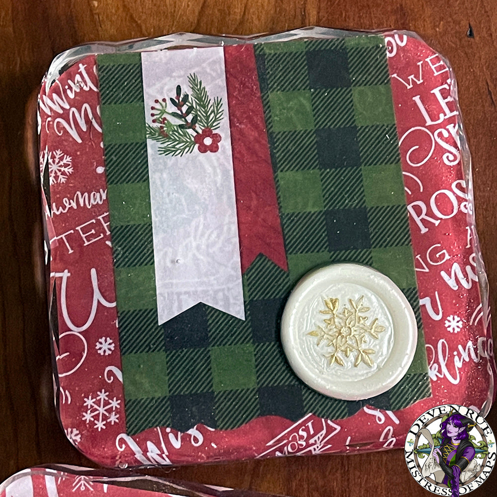 A close up of a resin coaster with green and black plaid, winter floral illustrations, and a white wax seal with gilded snowflake.