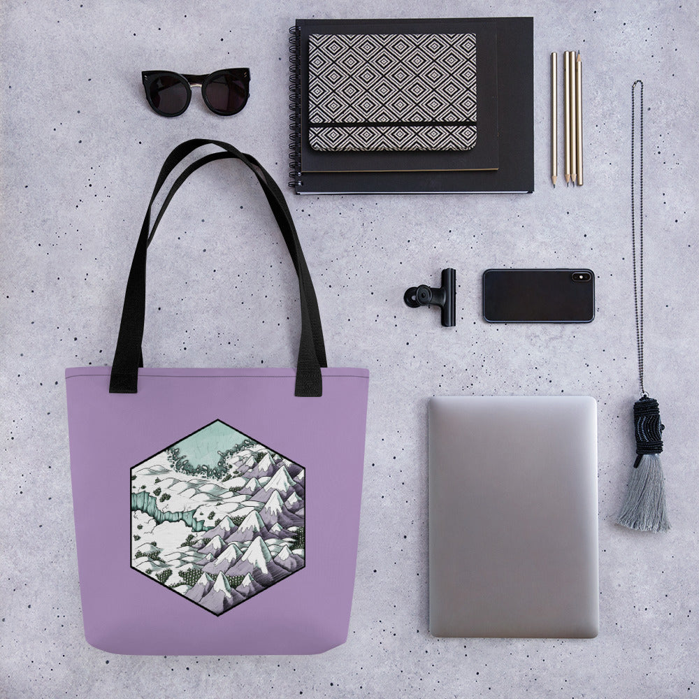 A hexagonal map illustration of a snowy, mountainous landscape on a lavender tote bag with common travel accessories for scale.