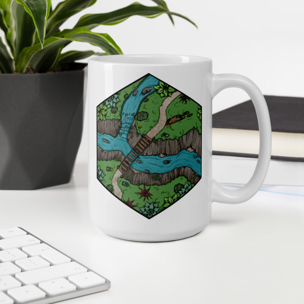 A mug with a hexagonal portion of a river crossing illustration sits near a plant, books, and a keyboard.