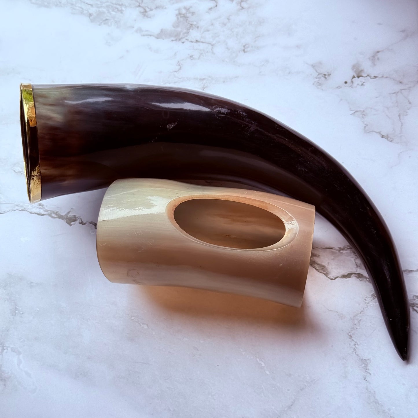 Horn shaped drinking vessel with gold trim along the mouth laying next to a lighter colored holder also made out of ox horn.