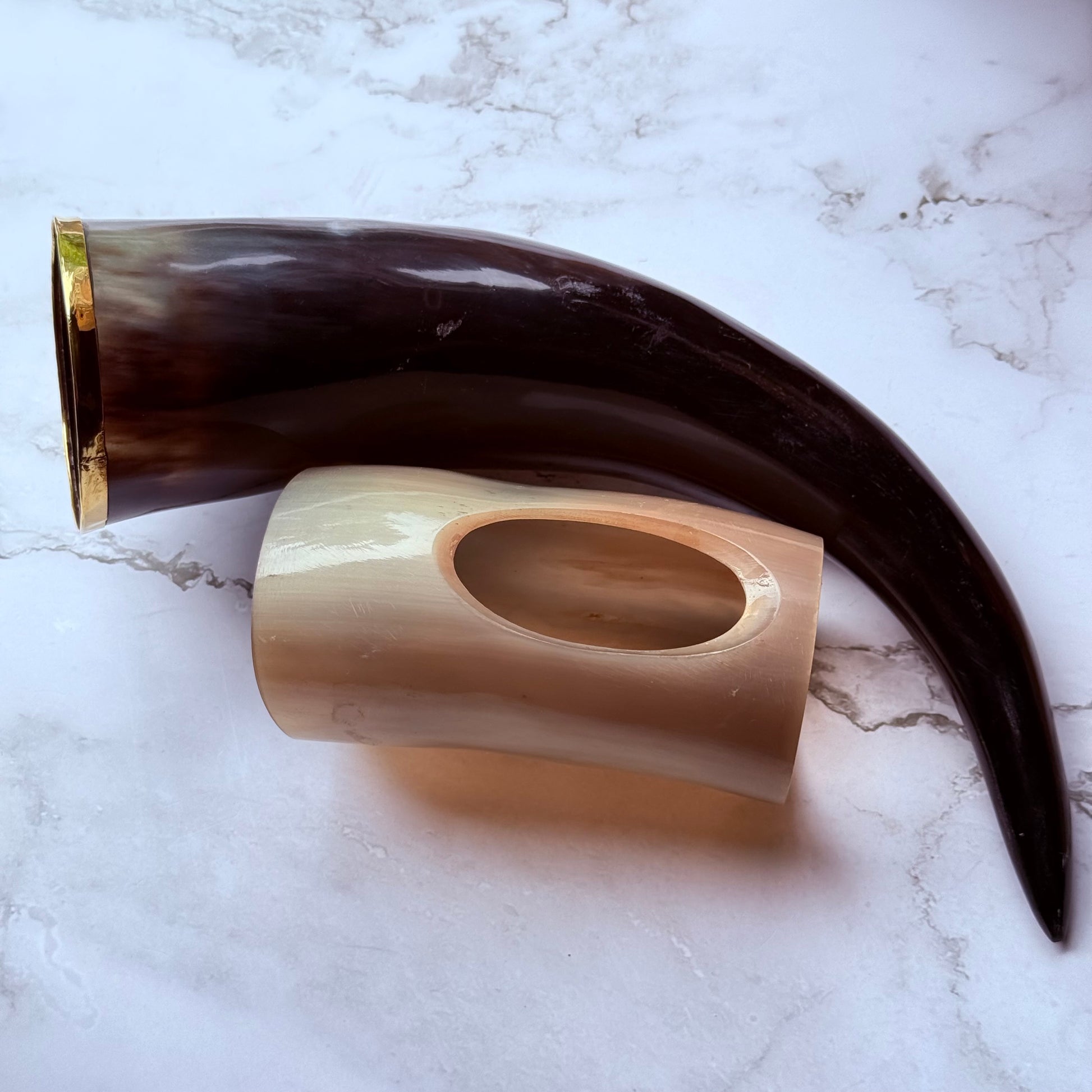 Horn shaped drinking vessel with gold trim along the mouth laying next to a lighter colored holder also made out of ox horn.