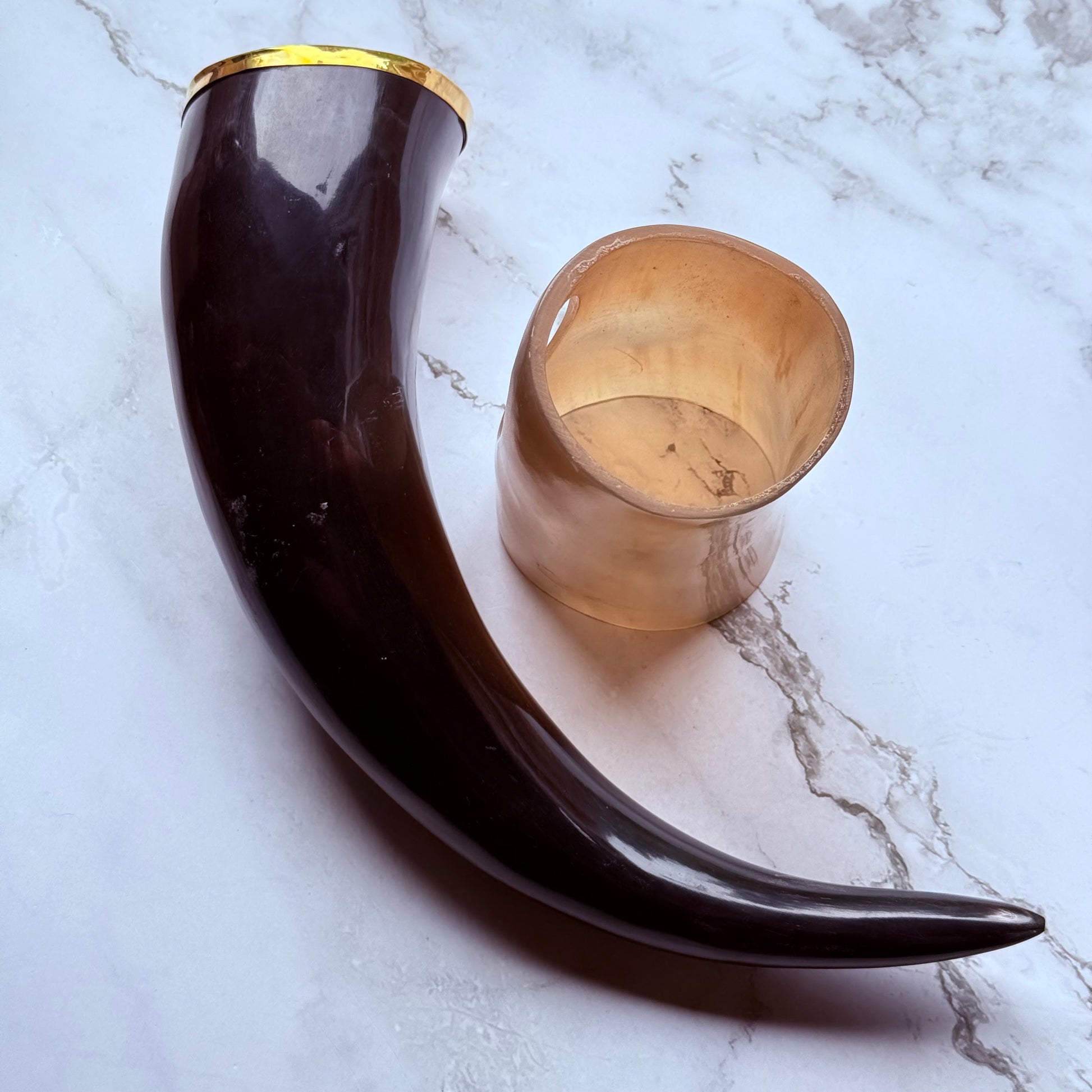 Horn shaped drinking vessel with gold trim along the mouth laying next to a lighter colored holder also made out of ox horn.
