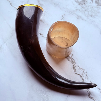 Horn shaped drinking vessel with gold trim along the mouth laying next to a lighter colored holder also made out of ox horn.