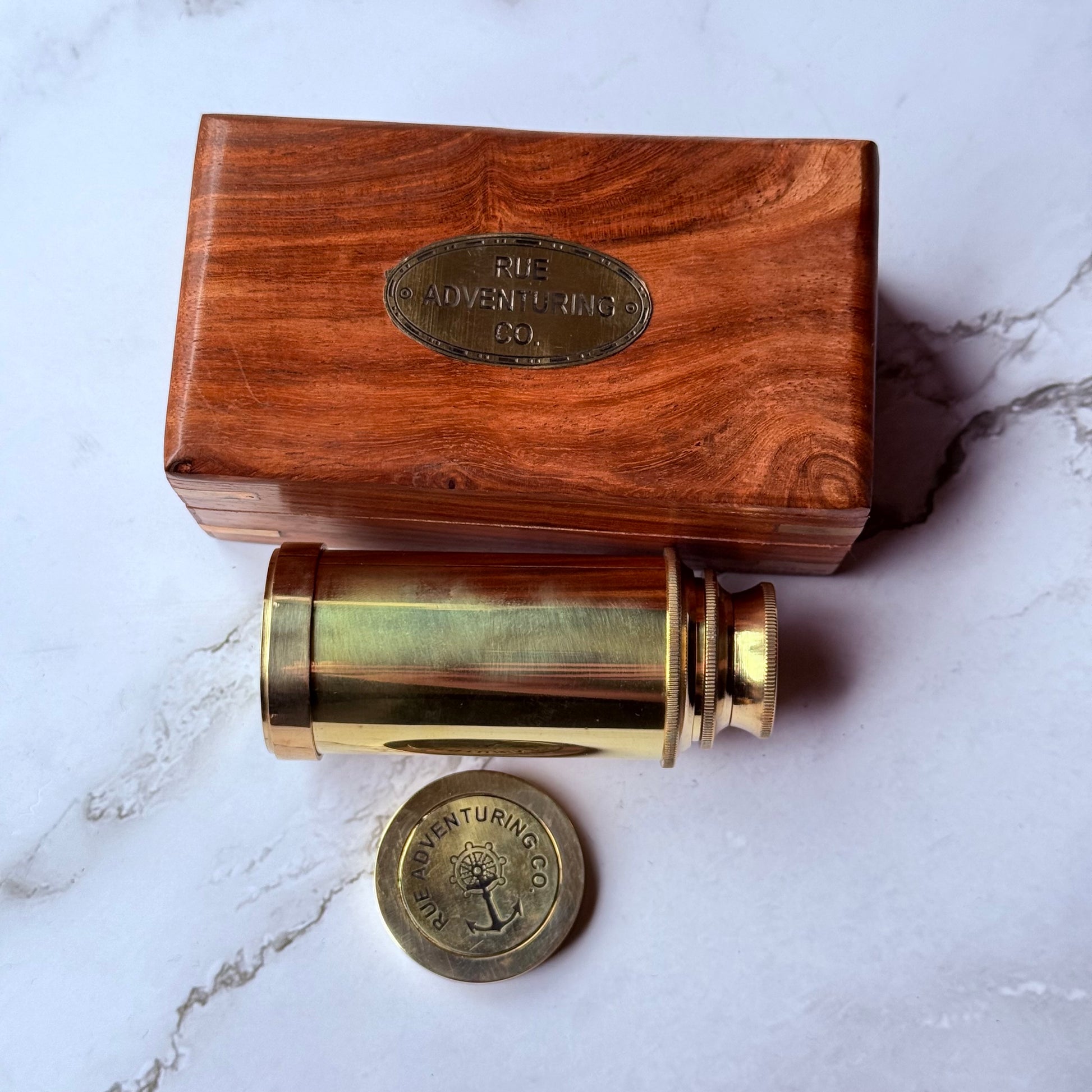 Brass telescope and wooden box with 'The Adventuring Co.' branding on a marble surface.