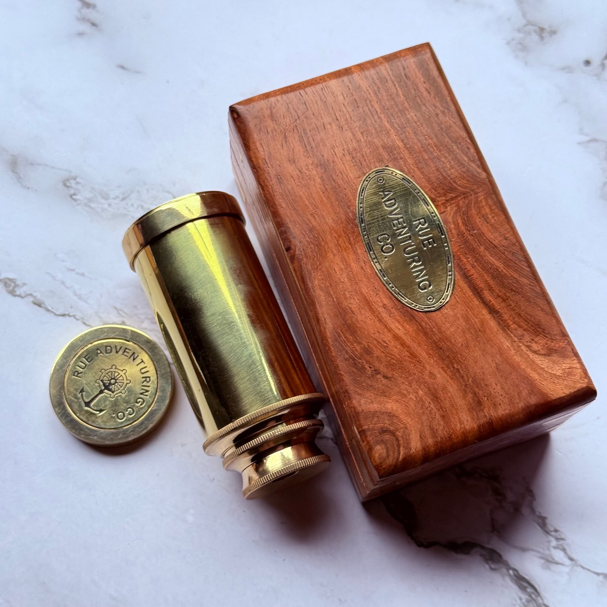 Brass telescope and wooden box with 'Rue Adventuring Co' branding on a marble surface