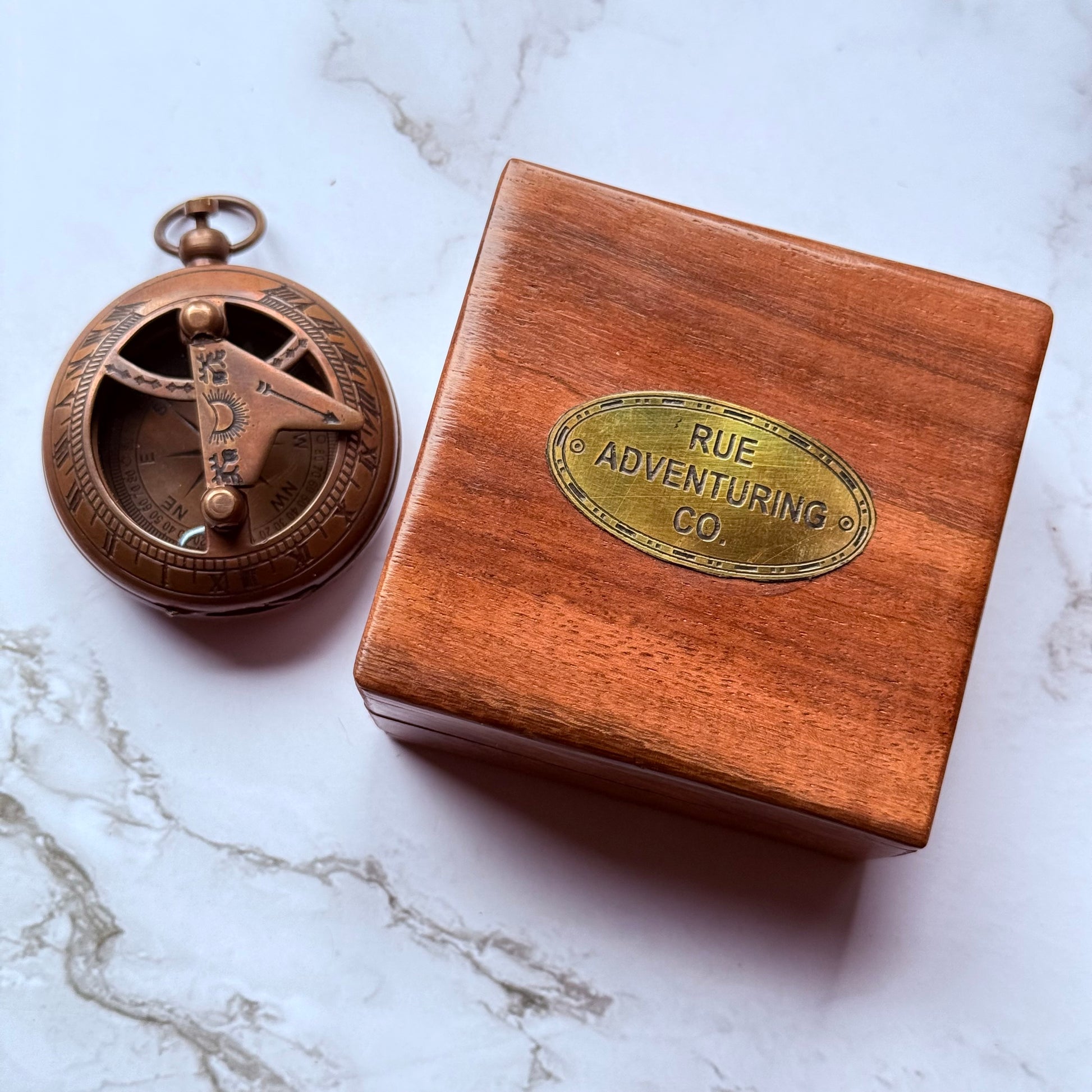 Bronze compass and wooden box with 'Rue Adventuring Co' on a marble surface