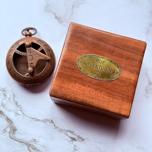 Bronze compass and wooden box with 'Rue Adventuring Co' on a marble surface
