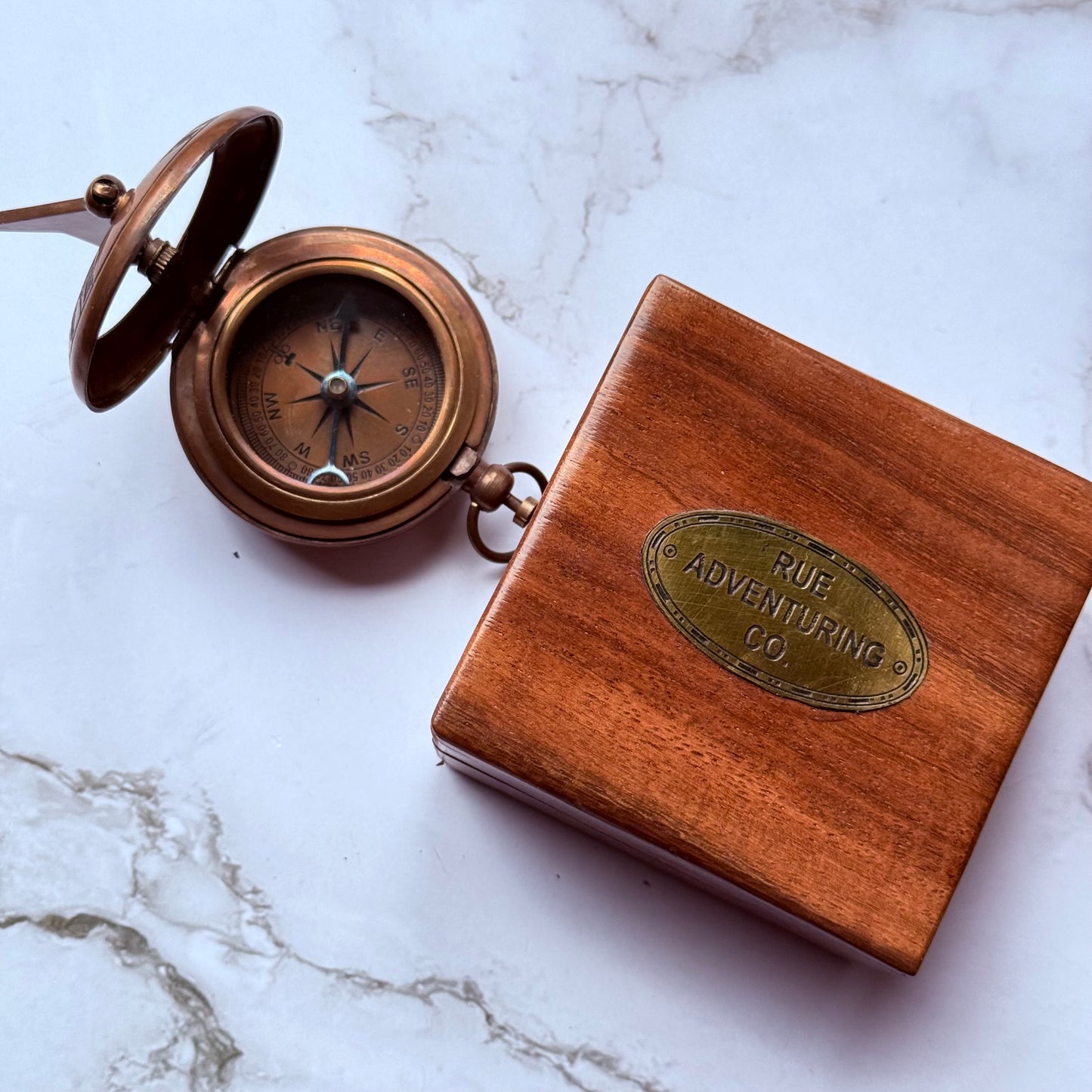 Bronze compass and wooden box with 'Rue Adventuring Co' emblem on a marble surface