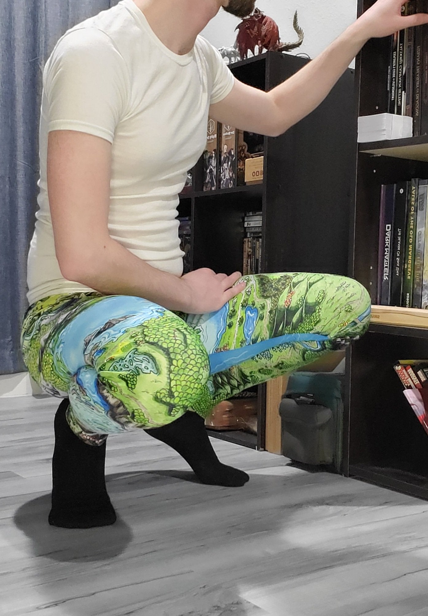 Josh Simons searches his bookshelf while crouching down in his Taur'Syldor map leggings by Deven Rue.