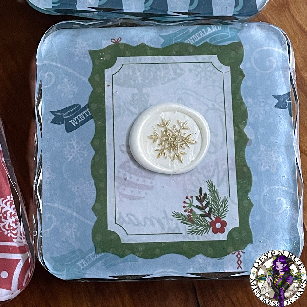 A close up of a resin coaster with a post card motif, winter floral illustration, and white gilded wax seal.