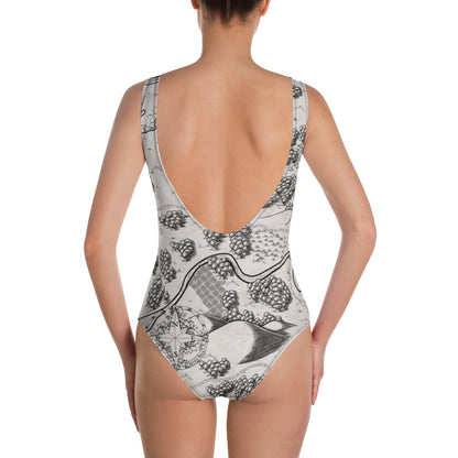 A model wears the Humble Beginnings map one piece swimsuit by Deven Rue, back view.