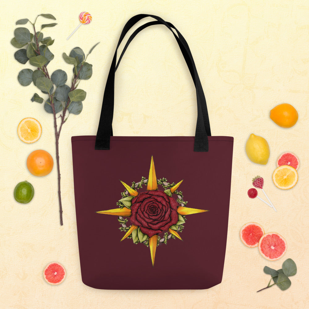 A burgundy tote bag with a compass rose illustration featuring a rose in the middle.