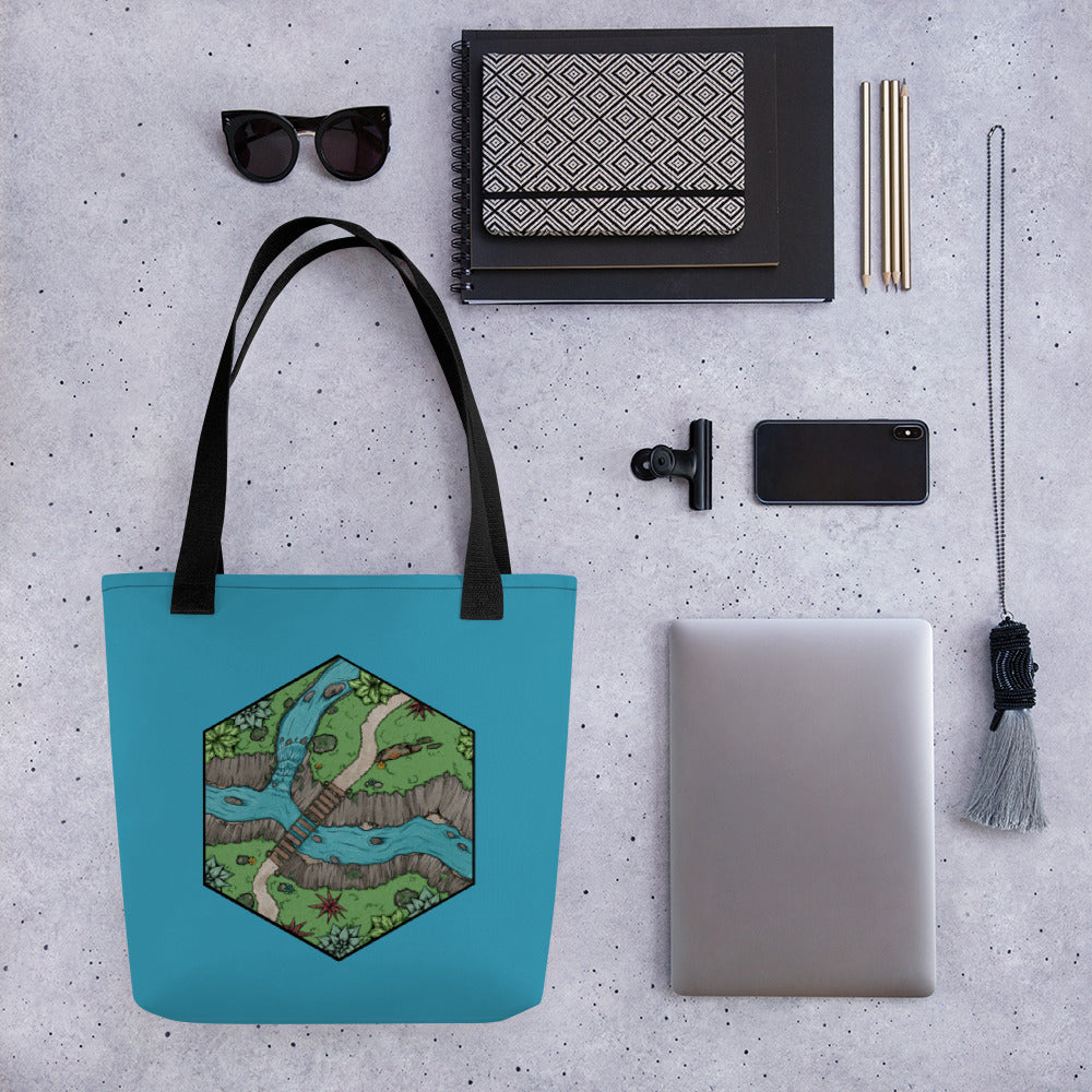 A tote bag in blue with a hexagonal portion of a river crossing map sits with common office items for scale.