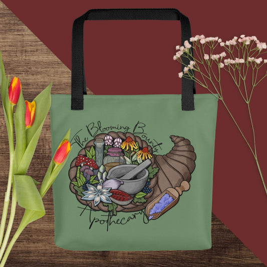 A sage green tote bag with The Blooming Bounty Apothecary logo featuring a cornucopia of herbalism supplies, sits with flowers.