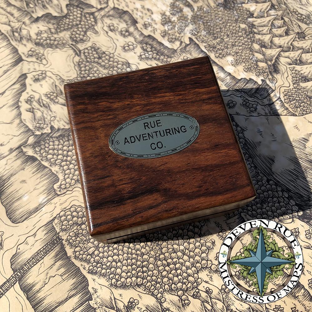 The compass and sundial in the Rue Adventuring Company branded box sits on a map.
