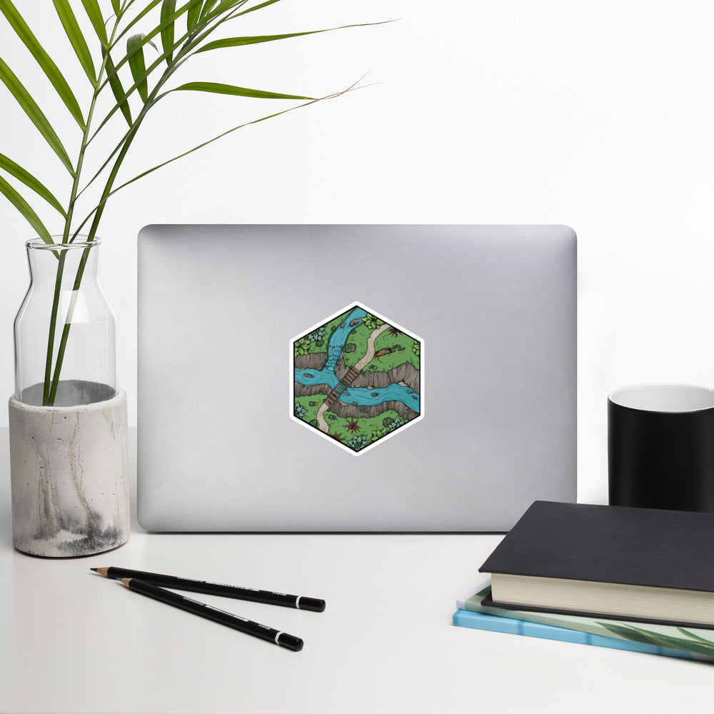 A sticker of a hexagonal portion of a river crossing map illustration is shown on a laptop with common office items for scale.