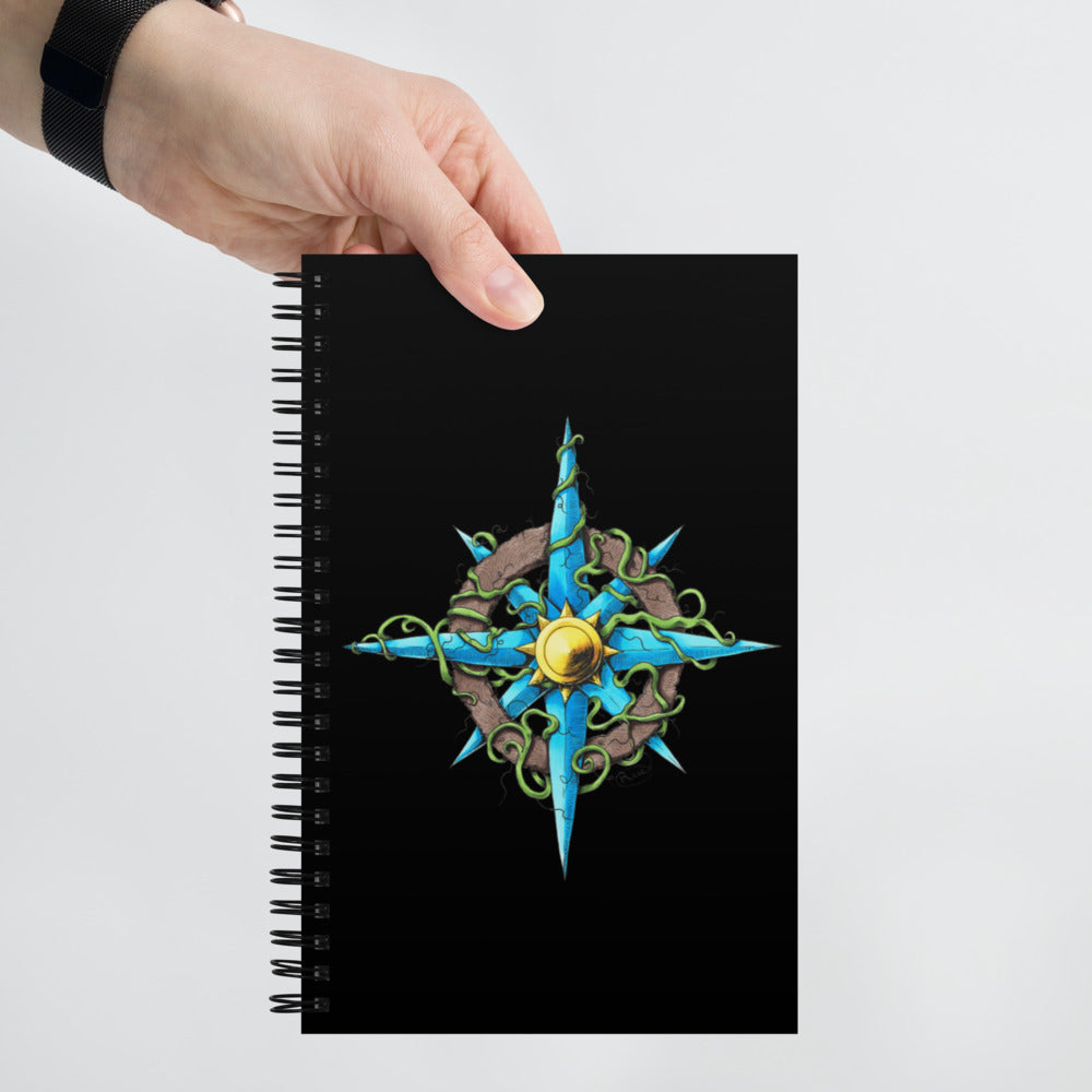 A hand holds a black spiral notebook with the Wilds Compass Rose illustration on the front.