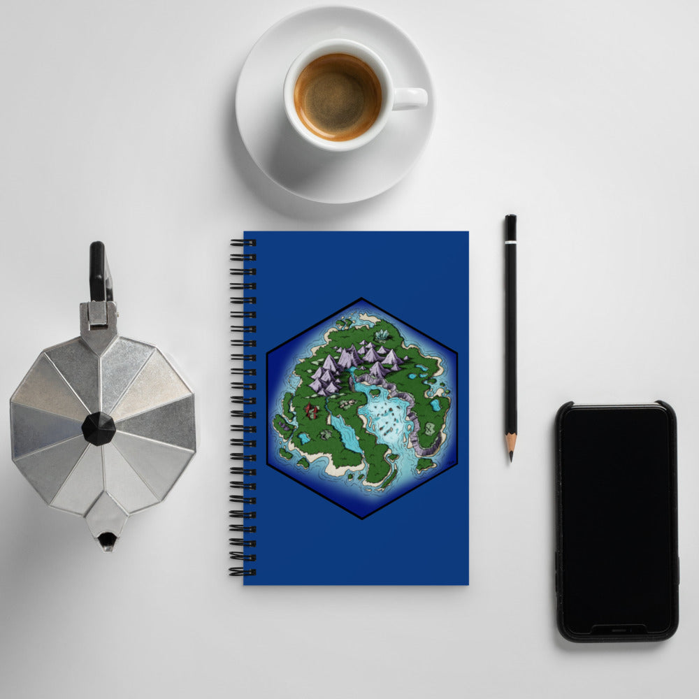 The Skycaller Island hexagonal map illustration is shown on the front of a royal blue spiral notebook surrounded by kettle, cup and saucer, pencil, and phone for scale.