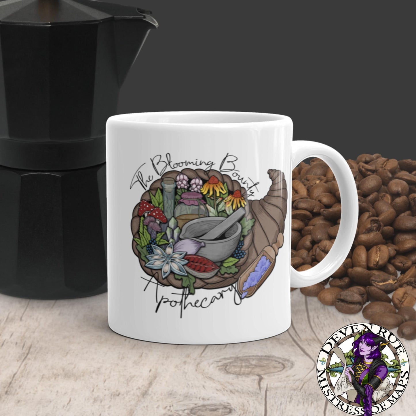 A white mug with The Blooming Bounty Apothecary logo featuring a cornucopia of herbalism supplies, sits with coffee beans and a kettle.