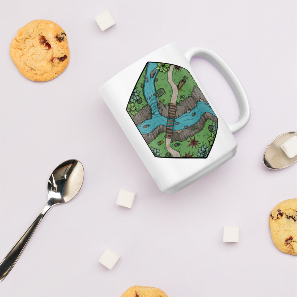A white mug with a hexagonal portion of a river crossing illustration sits surrounded by cookies, sugar cubes, and spoons to show size.